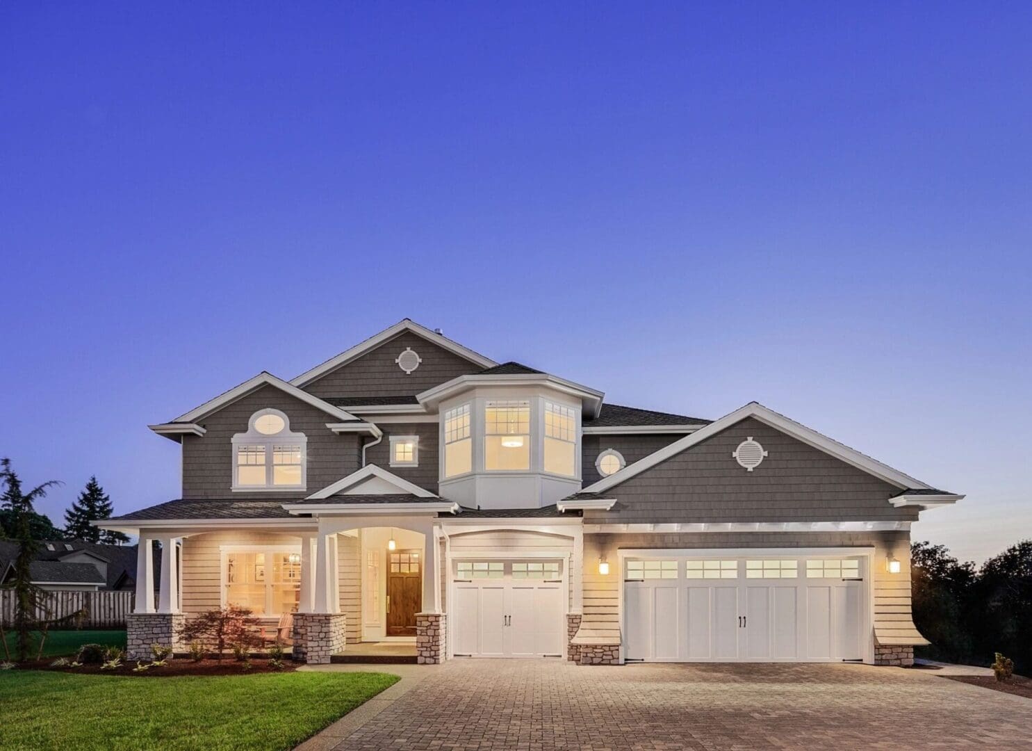 A large two-story house with illuminated windows, a grey exterior, a three-car garage, and a paved driveway, set against a clear evening sky.