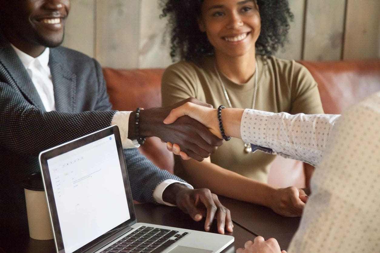 Two people are shaking hands over a table, while a laptop is open in front of them. Another person is sitting next to them, smiling.