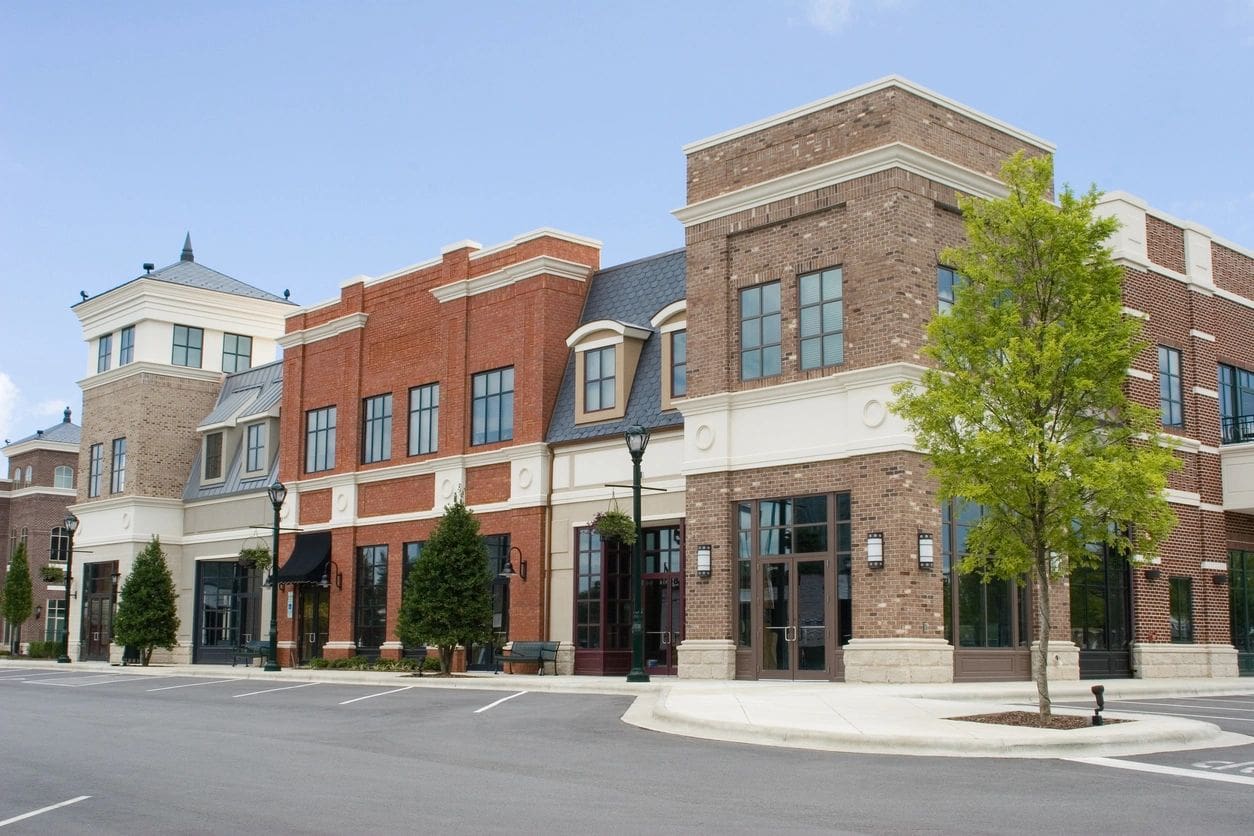 A modern commercial building with various storefronts, large windows, and a mix of brick and stucco siding, set on a quiet, empty street with a few small trees.